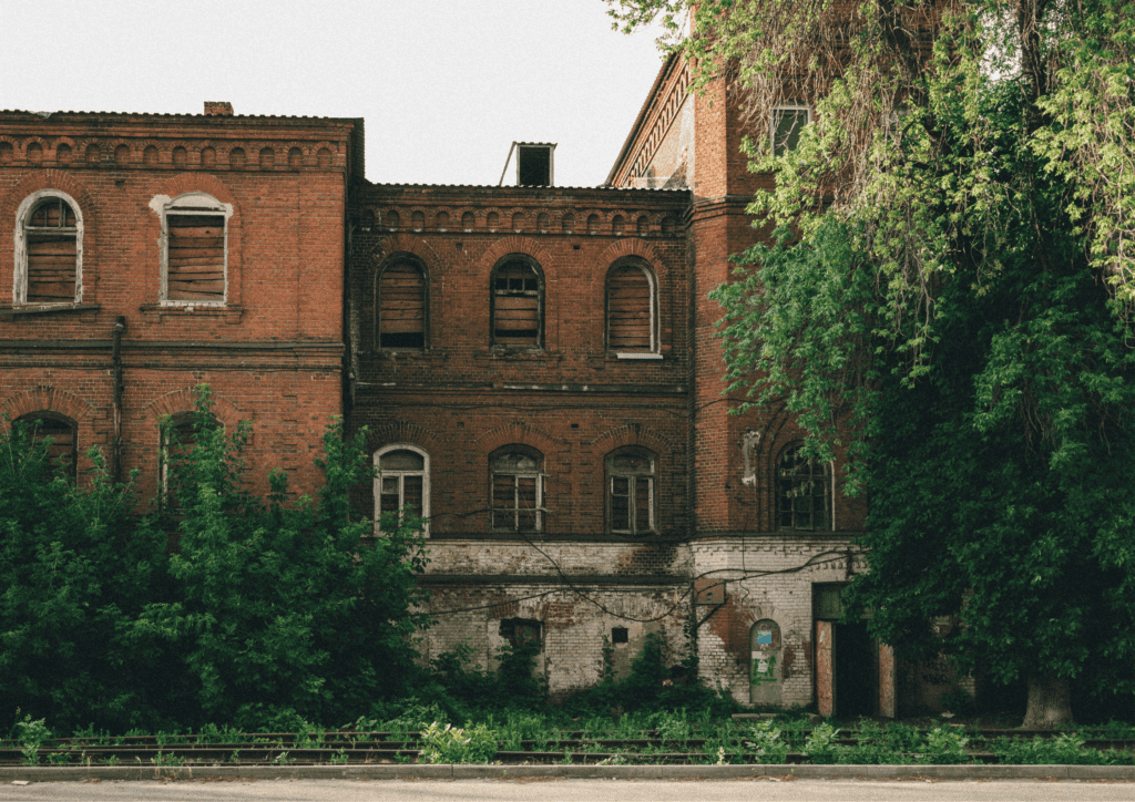 A large red brick building with boared up windows, signs of forced entry and damage as well as overgrown trees