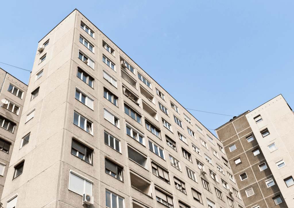 A block of grey flats with balconies inset into the building, shown as if looking up from the pavement below