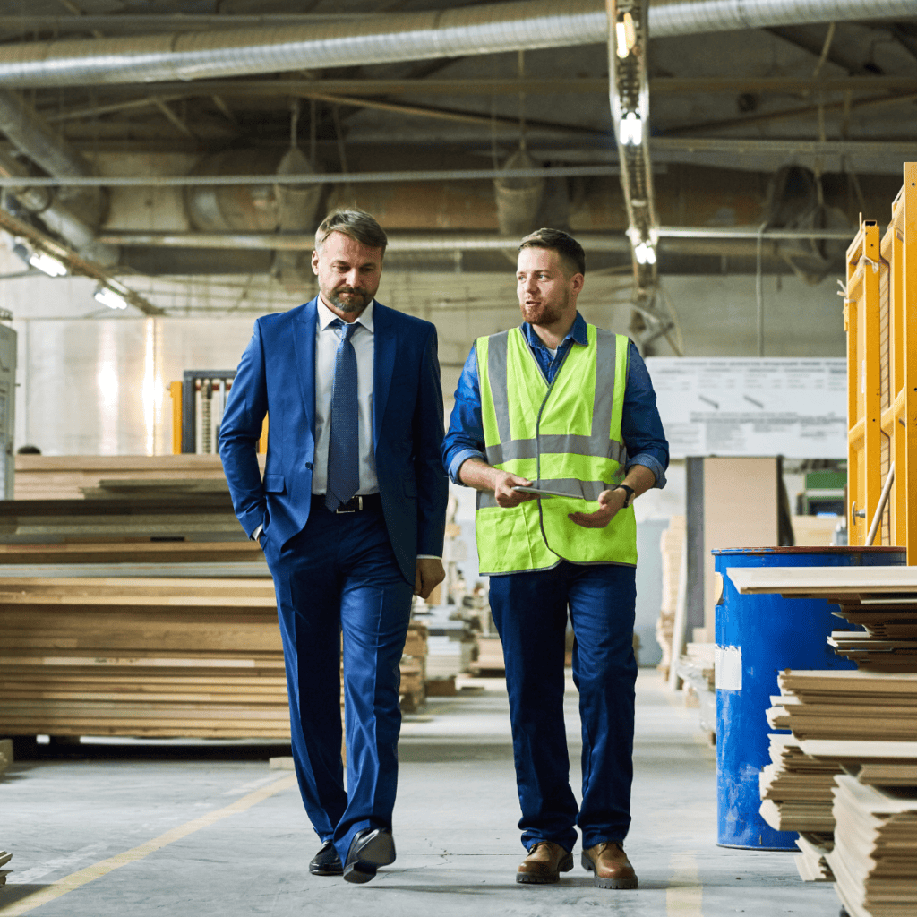 A man in a blue business suit walks with a contractor in a high visibility jacket, conversing and surrounded by building materials