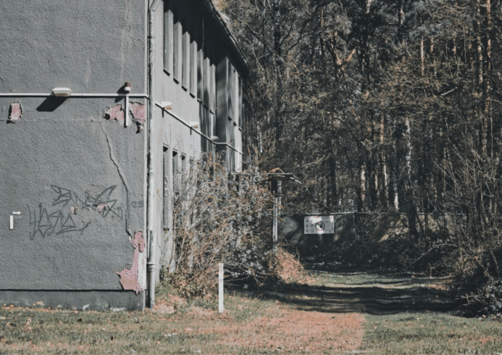 An abandoned building clad in grey sits off of the side of a dirt road, surrounded by trees. It has visible signs of decay.
