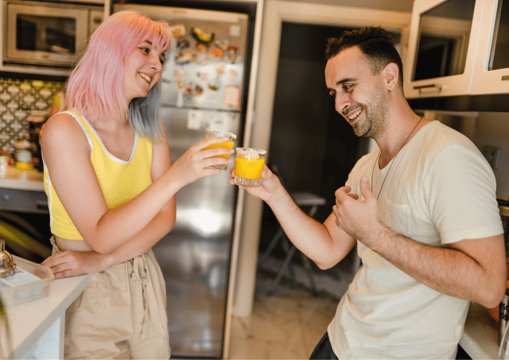 A woman with pink hair in a yellow tank top clinks a glass of orange juice with a man with short brown hair.