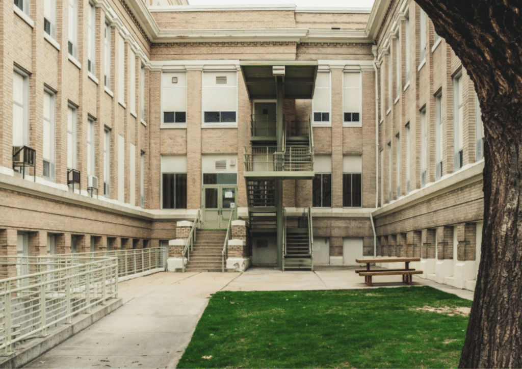 A courtyard with green grass, beige paving stones, surrounded by a beige panel tiled building with lots of windows.